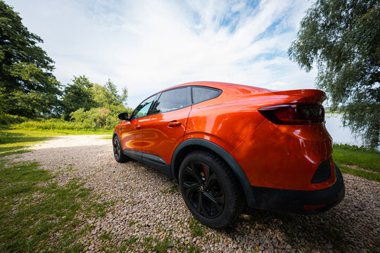 Vibrant orange Renault Arkana RS Line rests on a gravel pull off near water and trees, wide angle ground level view shows LED tail lamps, black wheels, red calipers.
