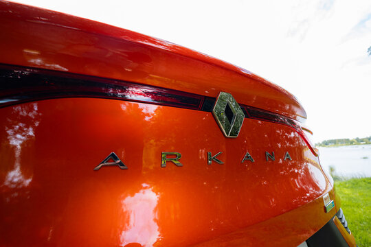 Rear hatch of Renault Arkana RS Line shows diamond badge and ARKANA text under a slim red light strip on vivid orange paint, daylight outdoor scene, low wide angle view.
