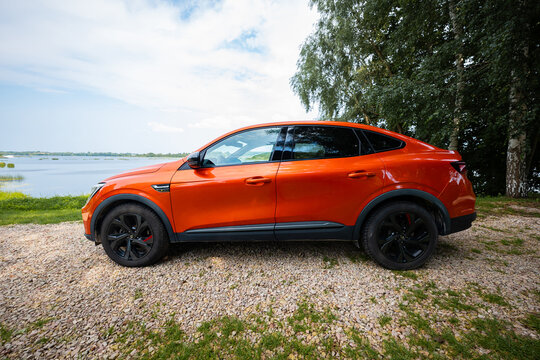 Renault Arkana RS Line in vibrant orange parks by a lakeside turnout. Side profile shows gloss black 5 spoke wheels, black mirrors, side badging, and a sloping roofline.