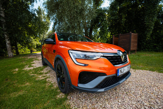 Vivid orange Renault Arkana RS Line parks three quarter front on gravel, low angle shows C shaped LED headlights, diamond badge, black alloys, and RS Line badges.