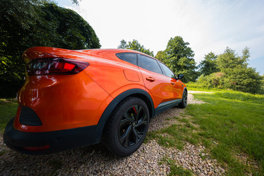 Orange Renault Arkana RS Line parks on a gravel path beside grass. Wide lens view shows tapered roofline, black roof spoiler, gloss black wheels, and RS Line badges.