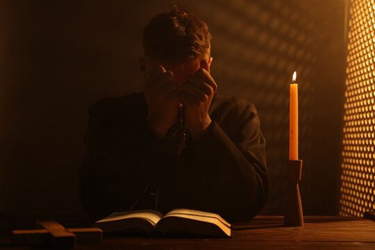 Priest praying with Holy Bible and rosary at wooden table in confessional booth