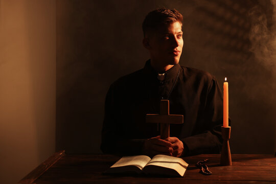 Priest holding cross at wooden table with Holy Bible, rosary and candle in confessional booth. Space for text