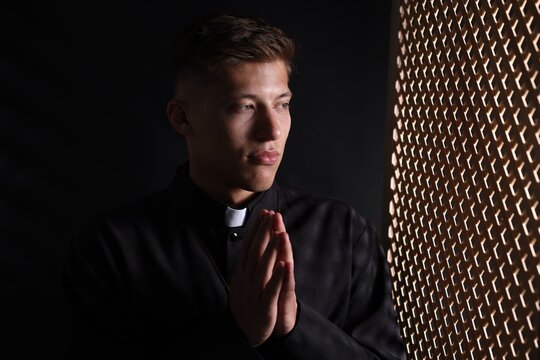Priest with clasped hands praying near partition in confessional booth