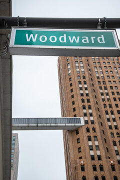 View of the Woodward Avenue street sign hanging above a city street with a tall historic brick building and a pedestrian skybridge in the background in Detroit, Michigan, United States.