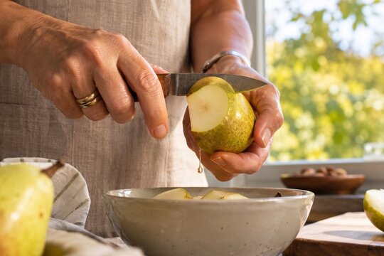 Person peeling a fresh pear with a knife over a bowl in the kitchen