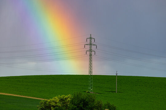 View of a rainbow arching over a green field with a power transmission tower and utility pole in Brno, South Moravian Region, Czechia.