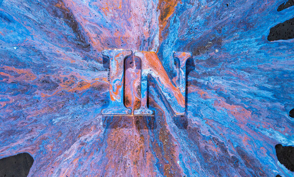 Metal uppercase IN coated in flowing blue, orange, and purple paint with radiating splash