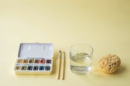 Watercolor paint set with brushes, a glass of water, and a sponge for artistic painting on a plain yellow background