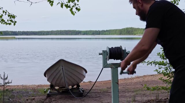 A man operates a manual hand winch to lower and release a rowing boat into the lake water from the shore. Horizontal.
