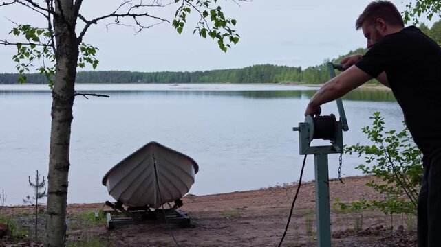 A man operates a manual hand winch mechanism to pull a rowing boat onto a lake shore on a peaceful summer day. Horizontal.