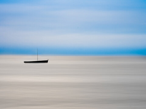 Lone sailboat drifts on calm, misty ocean waters under a soft blue sky