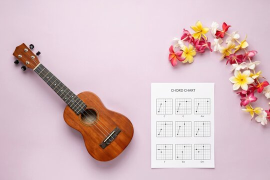 Wooden ukulele with musical chord chart sheet and tropical Hawaiian flowers on purple background