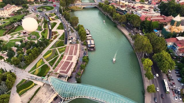 Panoramic drone shot looking down a city river flanked by a modern landscaped park with a moored hot air balloon and contemporary architecture on one side, and traditional city buildings 