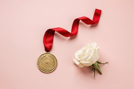 A single gold medal with a red ribbon lies next to a white rose, symbolizing achievement and celebration against a soft pink background