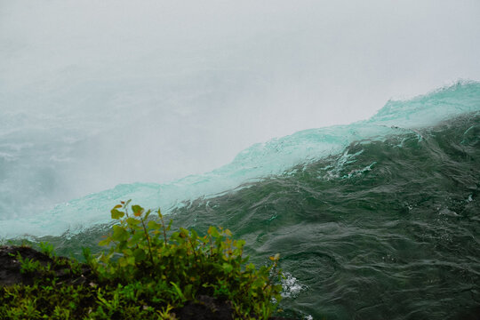 View of Niagara Falls with rushing turquoise water cascading over the edge and lush green plants on the rocky bank in Niagara Falls, Ontario, Canada.