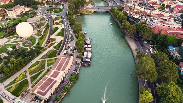 High-angle aerial drone shot showing a wide river with a motorboat cruising, flanked by a lush green city park, an iconic glass pedestrian bridge, a tethered helium balloon, and urban streets during t