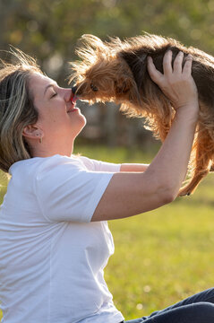 A young woman lifting a small dog up high, and the dog licking the woman's nose on a sunny day.