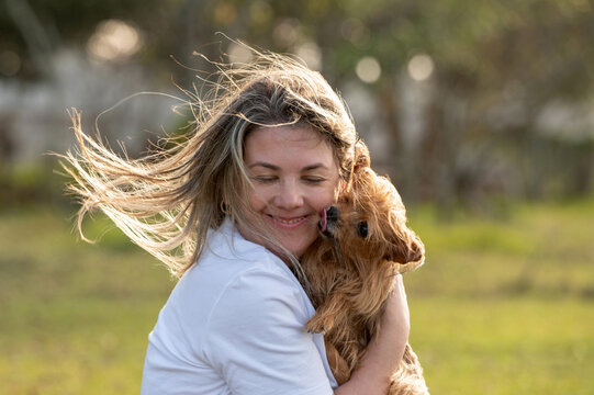 A young woman holding a small dog in her lap, eyes closed, with love; the dog is licking her face. Hair blowing in the wind. In a park, sunlight, summer day.