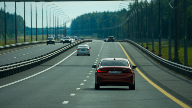Moscow, August 03 2026, Red car drives on a multi-lane highway. Other vehicles travel ahead in both directions. Tall poles line the road with green trees. Clear sky shines over the scene. Capture