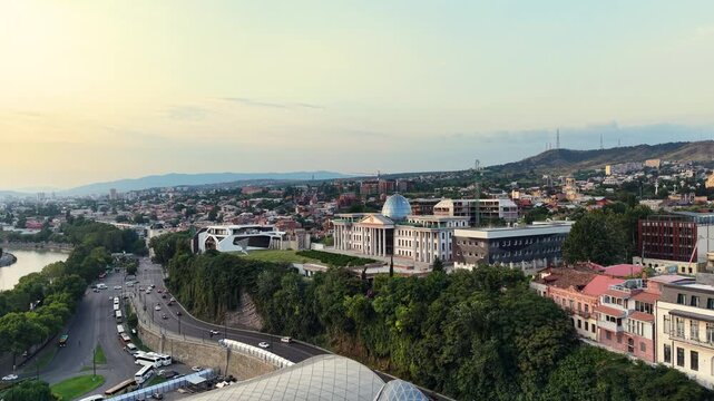 Wide aerial drone shot showcasing the classic Orbeliani Palace with its glass dome sitting on the hills of Avlabari district overlooking the city of Tbilisi and the Mtkvari river.