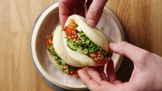 Spicy shrimp bao bun is shown in hand. This delicious shrimp bao bun is served with hot sauce, cucumber, scallion and sesame. Shrimp bao bun is Asian street food, seafood snack or takeaway lunch.