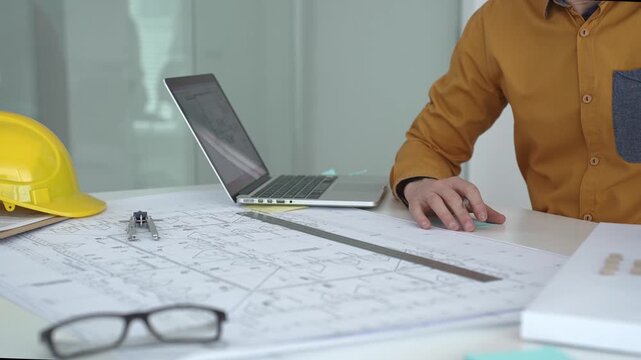 Engineer working on architectural blueprints and plans, typing on a laptop with a compass, ruler, safety helmet, glasses, and a building model on the desk