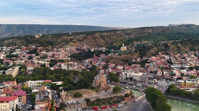 Scenic drone shot sweeping over historic Old Tbilisi in Georgia, showing Metekhi church on the cliff, traditional houses with red roofs, Narikala fortress on the hill, and the Kura river valley.
