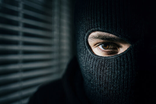 Burglar in black ski mask peeking through window blinds