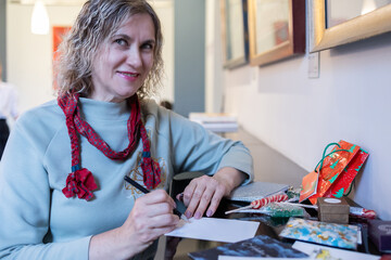 Preparing holiday postcards with drawings and decorations smiling woman crafting seasonal greetings cozy indoor scene. © lara-sh