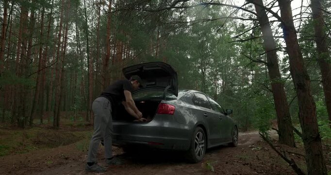 Man putting on jacket near open car trunk in rainy forest with audio included