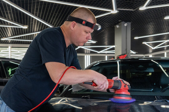 Professional auto detailer with a headlamp polishes a black vehicle hood using an electric dual action orbital polishing machine with a blue foam pad buffer tool in a car workshop garage. 