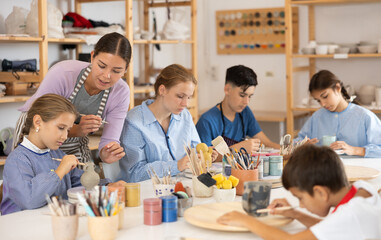 Fototapeta premium Teenagers and children at a ceramics workshop in an art studio. Woman teacher shows how to paint clay dishes with a brush