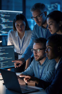 Diverse team of software developers huddle around laptop and monitor in dark office with blue light. People collaborate on code, discussing project strategy. They work together on digital innovation.
