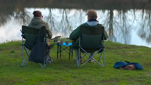 Couple enjoying hot drinks during outdoor camping trip. Rear view of a man and a woman sitting on folding chairs by a serene lake, pouring hot drinks from a thermos into mugs while enjoying a nature