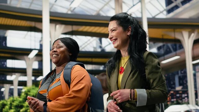 Happy women walk through the bus terminal while chatting, carrying backpacks in the shuttle station and enjoying a friendly conversation before departure.