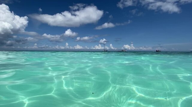 Breathtaking low-angle view of the crystal-clear turquoise and emerald waters in Zanzibar, featuring gentle sunlit ripples on the surface, distant boats, and fluffy white clouds under a brilliant blue