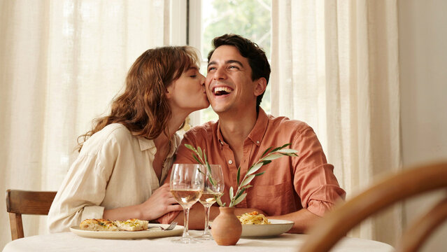 Affectionate young couple enjoying a romantic dinner date at home with wine with portrays young woman kissing her laughing boyfriend cheek during romantic meal sunlit dining area