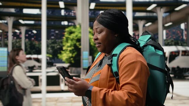 Smiling woman uses a smartphone travel app while walking through a bus terminal with a packed backpack; the passenger reads journey information on the phone screen before departure.