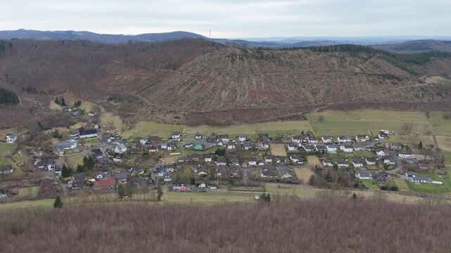 Aerial drone footage panning left over Grund village in a valley surrounded by hills, featuring a notable deforested hillside. Located in North Rhine-Westphalia, Germany.