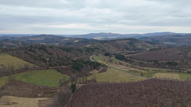 Boom up and push in on a deforested hilly landscape in North Rhine-Westphalia, Germany. Features winding roads, green fields, and a distant lake under an overcast sky.