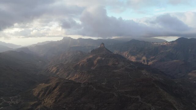 An aerial drone shot pushes in on a stunning, rugged volcanic landscape with Roque Bentayga mountain and winding roads on the Canary Islands, Spain. Capturing remote nature and tranquility.