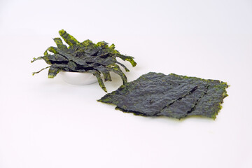 Fresh seaweed displayed on a clean white surface. One pile rests in a small bowl, slightly curled. Another lies flat, dark green with visible texture. Simple background highlights natural colors © Nurma