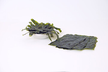 Fresh seaweed displayed on a clean white surface. One pile rests in a small bowl, slightly curled. Another lies flat, dark green with visible texture. Simple background highlights natural colors © Nurma