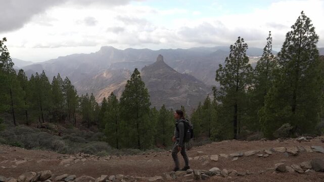 A lone hiker navigates a rugged trail on Gran Canaria, Canary Islands, with stunning views of the iconic Roque Bentayga formation and the vast volcanic landscape under a cloudy sky.