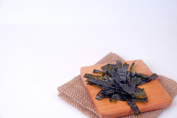 Dried seaweed rests on a wooden cutting board. Board sits atop a folded burlap napkin. White background offers clean, minimalist presentation. Seaweed leaves display natural texture and rich color © Nurma