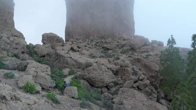 A hiker navigates a rugged, rocky trail to the majestic Roque Nublo monolith on the Canary Islands. The landscape is shrouded in atmospheric fog, creating a remote and adventurous scene.