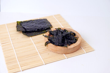 Kelp sheets rest on bamboo mat beside wooden bowl. Dried seaweed displays rich dark green hue. Simple background highlights natural textures and colors. Ideal for health, wellness © Nurma