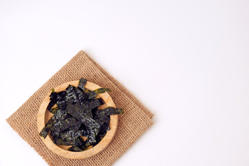 Dried seaweed rests in a wooden bowl. Burlap cloth provides rustic contrast beneath. Clean white background emphasizes simplicity and texture © Nurma