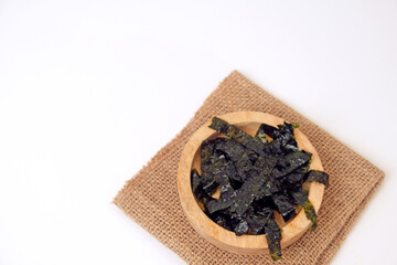 Dried seaweed rests in a wooden bowl. Burlap cloth provides rustic contrast beneath. Clean white background emphasizes simplicity and texture © Nurma